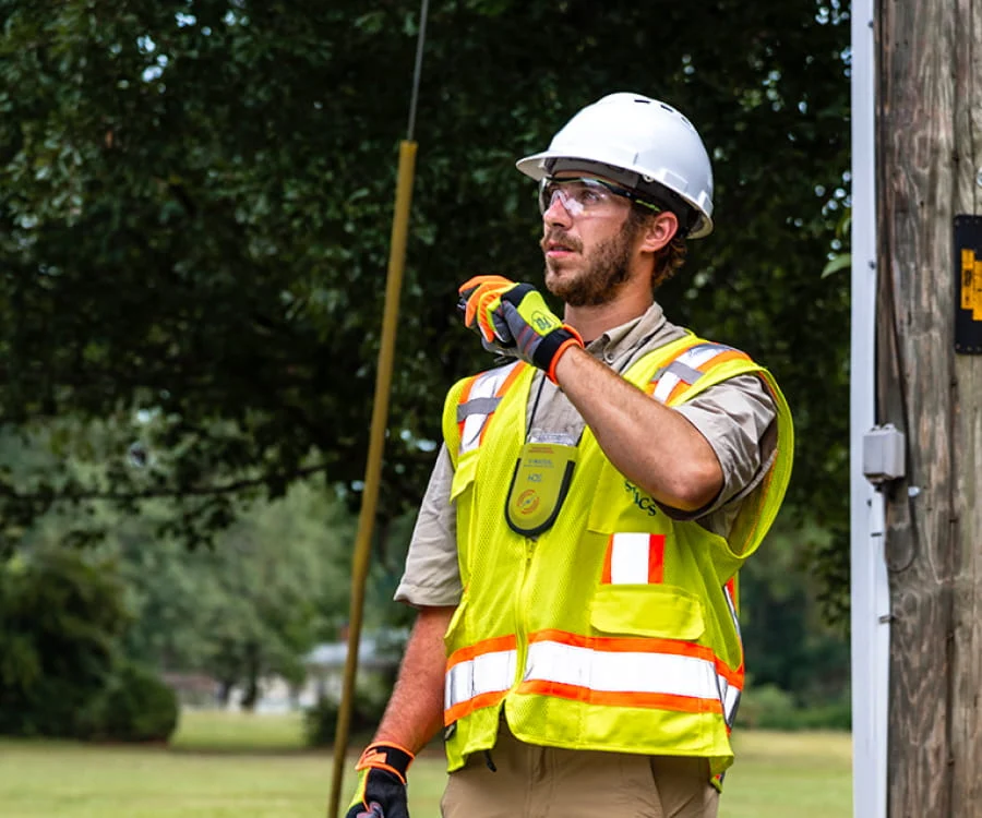 A DRG employee in a yellow vest and hard hat speaks into a walkie-talkie from a disaster site.